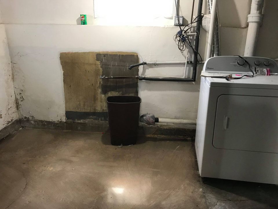 Basement laundry area with a dryer, exposed pipes and wiring, a stained wall section, and a trash can on a concrete floor.